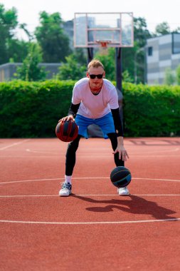 Uzun boylu, iki toplu bir basketbolcu parkta basketbol kortunda çalışırken salya sürme yeteneklerini sergiliyor.
