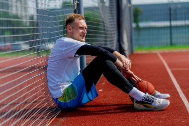 Elinde top olan uzun boylu bir basketbolcu parkta antrenman yaparken bir basketbol sahasının ağına yaslanır.