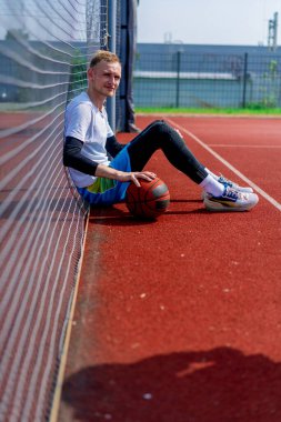 Elinde top olan uzun boylu bir basketbolcu parkta antrenman yaparken bir basketbol sahasının ağına yaslanır.