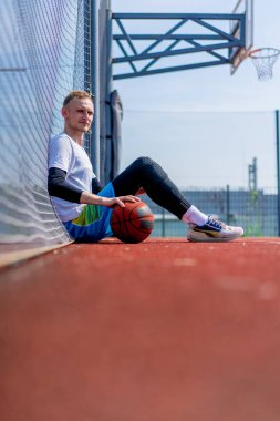 Elinde top olan uzun boylu bir basketbolcu parkta antrenman yaparken bir basketbol sahasının ağına yaslanır.