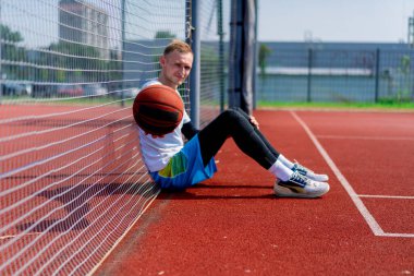 Elinde top olan uzun boylu bir basketbolcu parkta antrenman yaparken bir basketbol sahasının ağına yaslanır.