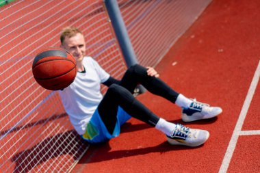 Elinde top olan uzun boylu bir basketbolcu parkta antrenman yaparken bir basketbol sahasının ağına yaslanır.