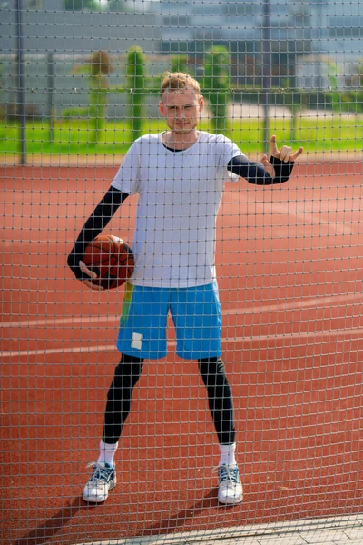 Tall guy basketball player standing on a basketball court on the street with ball in hand view through the fence netting