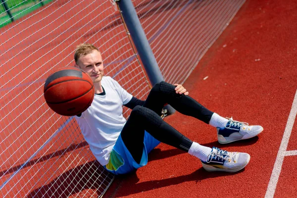 一位高个子篮球运动员在公园练习时 手里拿着一个球坐在篮球场的网子上 — 图库照片