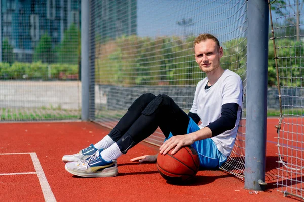 彼の手のボールを持つ背の高い男のバスケットボール選手は 公園での練習中にバスケットボールコートのネットに傾いています — ストック写真
