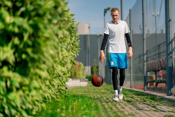 Uzun boylu basketbolcu, parktan basketbol sahasına doğru yürüyor ve topu sürüş yeteneklerini sergileyerek sürüyor.