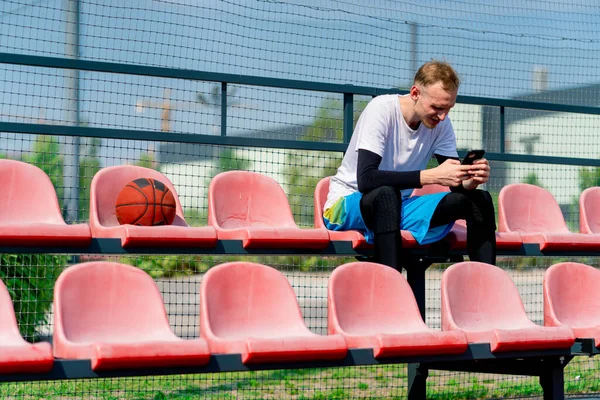 Wysoki Koszykarz Siedzi Wpatrując Się Smartfona Trybunach Boiska Kosza Obok — Zdjęcie stockowe