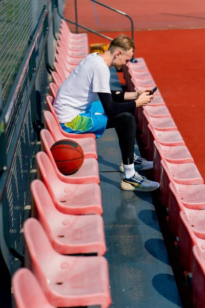 Vysoký Chlap Basketbalista Sedí Zírá Smartphonu Tribunách Basketbalového Hřiště Vedle — Stock fotografie