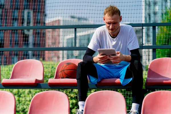 A tall guy basketball player sits looking at a tablet in the bleachers of a basketball court with ball next to him