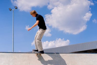 Genç bir kaykaycı şehir kaykay parkında gökyüzü ve bulutlara karşı bir buz pateni pistinin kenarında gösteri yapıyor.