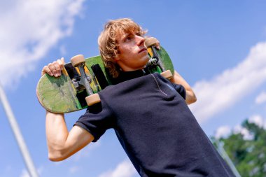 Paten parkında ve gökyüzünde omuzlarında kaykay tutan uzun saçlı genç bir kaykaycının portresi