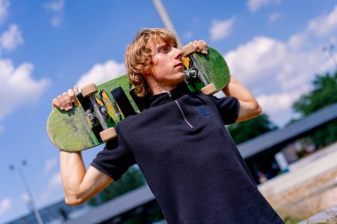 Paten parkında ve gökyüzünde omuzlarında kaykay tutan uzun saçlı genç bir kaykaycının portresi