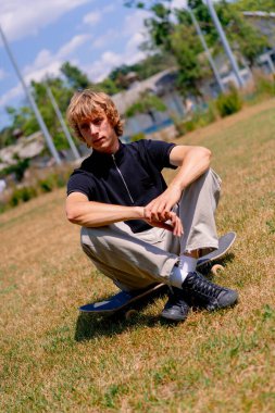 Genç ciddi kaykaycı, uzun saçlı kaykaycı çimlerin ve gökyüzünün arka planında oturuyor.