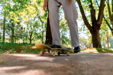 Genç kaykaycı kaykayla şehir parkının yolunda ağaçların ve gökyüzünün arka planına doğru gidiyor.