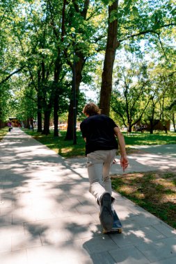 Genç kaykaycı kaykayla şehir parkının yolunda ağaçların ve gökyüzünün arka planına doğru gidiyor.