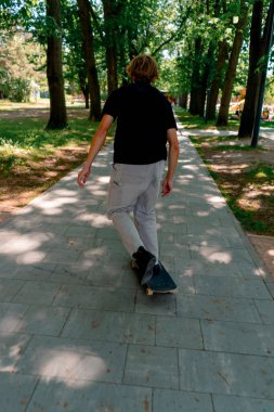 Genç kaykaycı kaykayla şehir parkının yolunda ağaçların ve gökyüzünün arka planına doğru gidiyor.