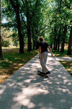Genç kaykaycı kaykayla şehir parkının yolunda ağaçların ve gökyüzünün arka planına doğru gidiyor.