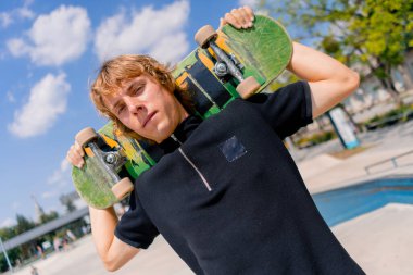 Paten parkında ve gökyüzünde omuzlarında kaykay tutan uzun saçlı genç bir kaykaycının portresi