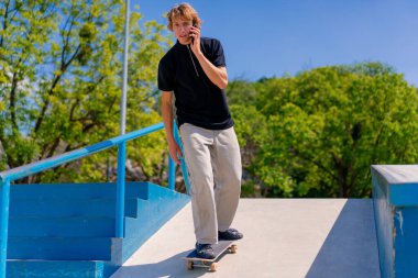 Şehirdeki kaykay pistinde uzun saçlı genç kaykaycı telefonda konuşuyor.