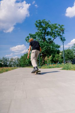 Genç kaykaycı kaykayla şehir parkının yolunda ağaçların ve gökyüzünün arka planına doğru gidiyor.