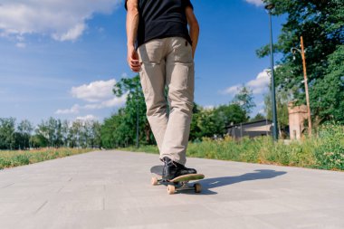 Genç kaykaycı kaykayla şehir parkının yolunda ağaçların ve gökyüzünün arka planına doğru gidiyor.