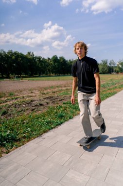 Genç kaykaycı kaykayla şehir parkının yolunda ağaçların ve gökyüzünün arka planına doğru gidiyor.