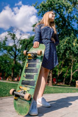 Ağaçların ve gökyüzünün arka planına karşı elinde kaykayla gezen elbiseli genç, gülen bir kaykaycı kız.