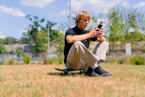長髪の若い深刻なスケーターの男は 芝生と空の背景に対して携帯電話を見ているスケートボードに座っています — ストック写真