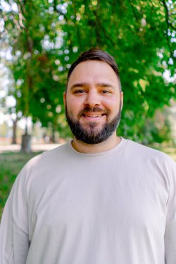 Beyaz tişörtlü, sakallı gülümseyen bir adamın portresi. Parkta, ağaçların arka planında duruyor.