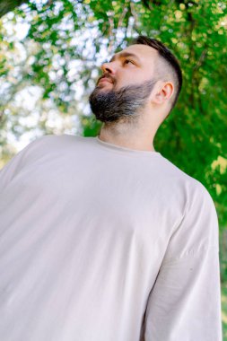 Beyaz tişörtlü, sakallı gülümseyen bir adamın portresi. Parkta, ağaçların arka planında duruyor.