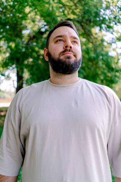 木の背景とサイズのモデルに対して公園に立っている白いTシャツのひげを持つ笑顔の男の肖像 — ストック写真