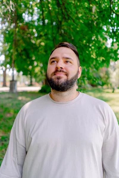 木の背景とサイズのモデルに対して公園に立っている白いTシャツのひげを持つ笑顔の男の肖像 — ストック写真