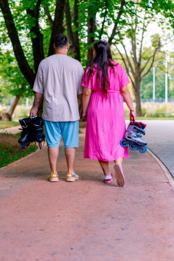 Genç bir çift, el ele tutuşup, büyük beden modellerin ellerinde patenleri tutarken parkta yürüyor.