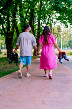 Genç bir çift, el ele tutuşup, büyük beden modellerin ellerinde patenleri tutarken parkta yürüyor.