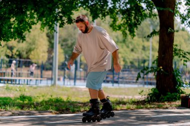 Sakallı genç adam şehir parkında paten kaymayı öğreniyor. Yeni şeyler öğrenmek ve vücut pozitifliği.