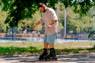 Sakallı genç adam şehir parkında paten kaymayı öğreniyor. Yeni şeyler öğrenmek ve vücut pozitifliği.