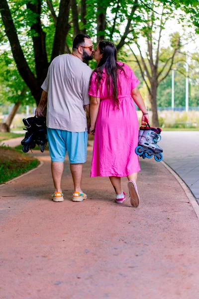 若いカップルは 手を握り プラスサイズのモデルの手にローラーブレードを保持しながら 公園を歩いています — ストック写真