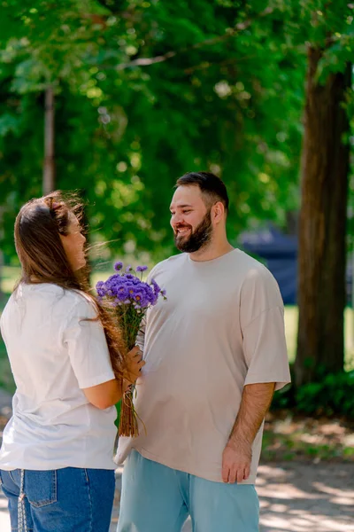 若いカップル陽気な男は木の背景とサイズのモデルとの日付の間に公園で女の子に花を与える — ストック写真