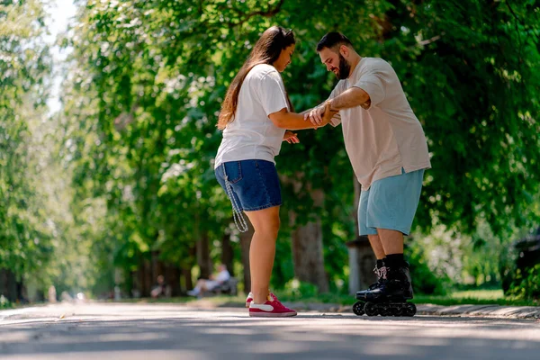 Young couple girl holds hands of the guy who is roller skating and helps him to learn to skate in park plus size models