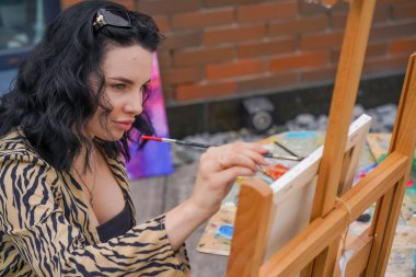 Genç ve güzel bir sanatçı sokakta bir sandalyeye oturur ve bir fırçayla sehpanın üzerinde duran bir resim çizer.