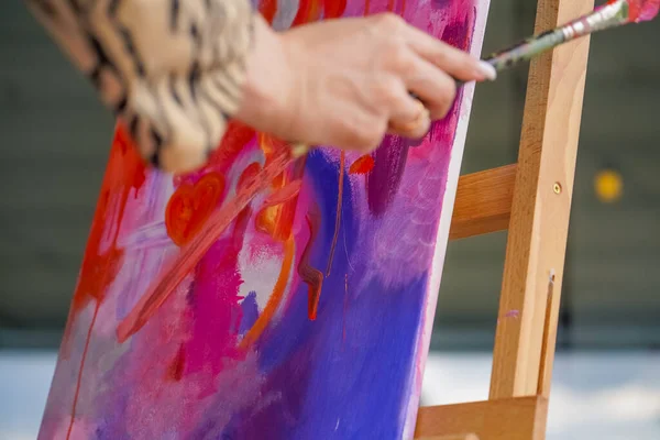 Close-up of a girl artist with a brush applying oil paint to a painting standing on easel in a studio for painting