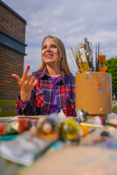 Portrait of cheerful girl artist finger painting paint on her face sitting at a painting table with tubes of paint and brushes on it