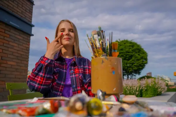 Portrait of cheerful girl artist finger painting paint on her face sitting at a painting table with tubes of paint and brushes on it