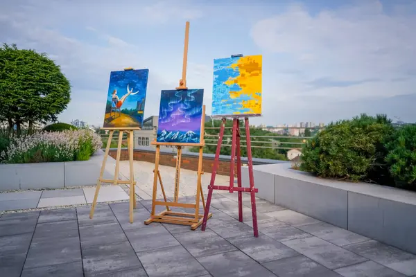 Three paintings stand on easels against a city backdrop in painting studio