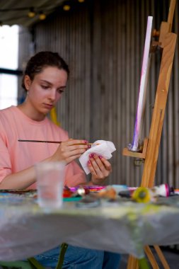 Sorunlu ciltli genç bir kadın sanatçı resim çizerken sehpasının yanına oturur ve boya fırçasını peçeteyle siler.