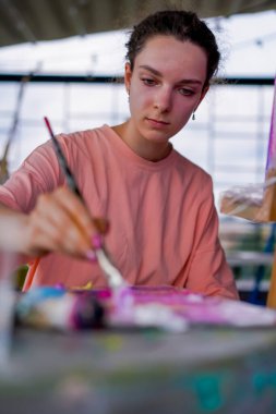 Sorunlu ciltli genç bir kadın sanatçı bir sehpanın yanında oturur ve paletin boyasını alır.