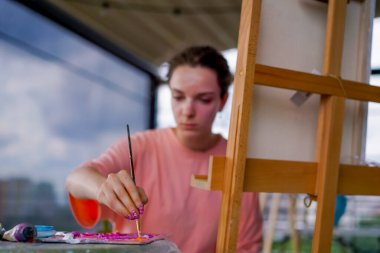 Sorunlu ciltli genç bir kadın sanatçı bir sehpanın yanında oturur ve paletin boyasını alır.