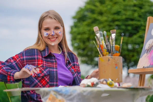 Portrait of a cheerful girl artist with oil paint on her face and brushes in her hands smiling against city background