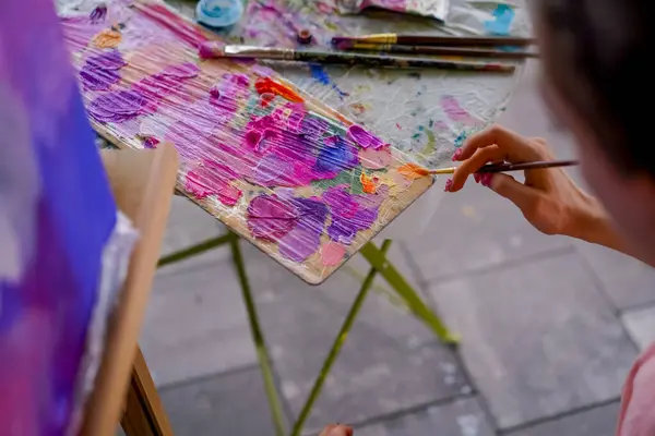 A close-up of girl artist sitting near an easel and taking paint from a palette with a brush the concept of love of fine art