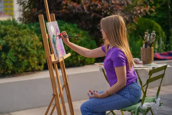 A young girl artist paints a picture with a brush on a canvas that stands on easel view from behind the back